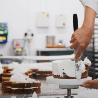Applying white cream frosting to a cake layer on a rotating stand in a professional bakery production area
